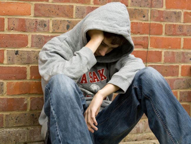 Boy wearing grey sweatshirt and jean with hood pulled over face sitting against a red brick wall