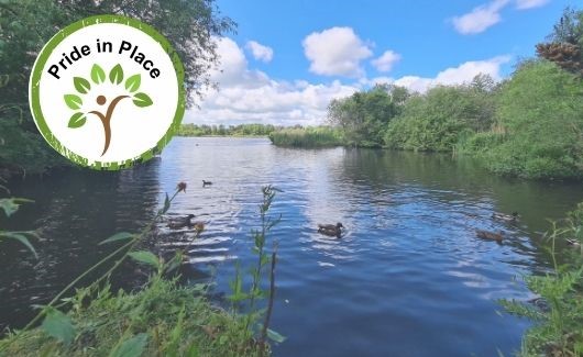 A still lake with ducks swimming surrounded by green trees and bushes . A Pride in Place logo of stylised tree with person underneath