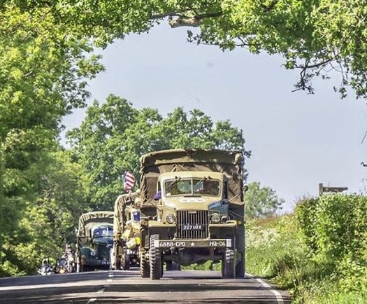 An older military truck leading a procession of classic miliary vehicles along a road with trees and hedges