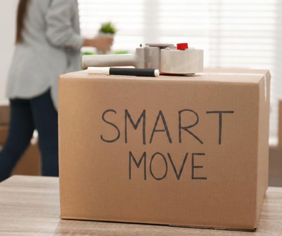 A cardboard moving box has the text "Smart Move" written on it in felt tip. A box sealer is visible on top of the box. A woman moves house plants in the background.