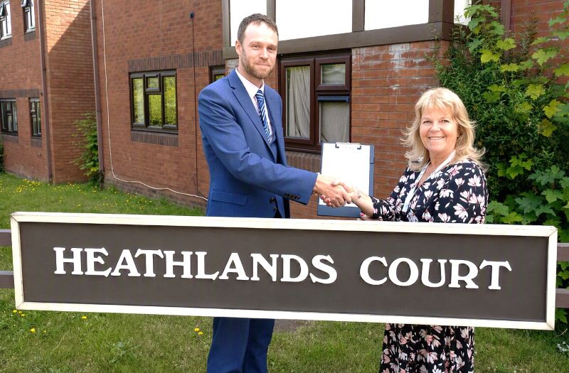 A man and a woman standing in front of a sign that says Heathlands Court, holdng between them a white document