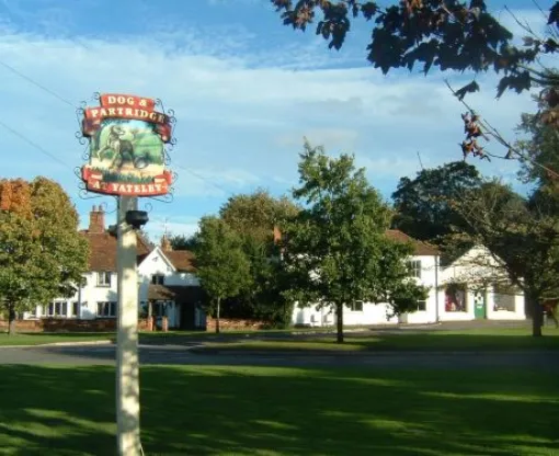 A quaint village scene featuring the "Dog and Partridge" pub sign in the foreground, with a traditional white building surrounded by lush greenery under a clear blue sky.