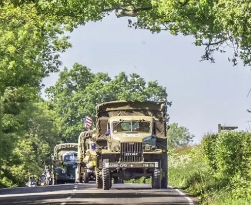 An older military truck leading a procession of classic miliary vehicles along a road with trees and hedges