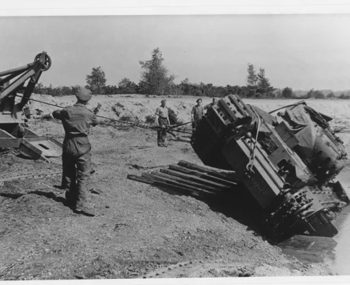 An overturned Churchill tank is being winched out of a pool of muddy water