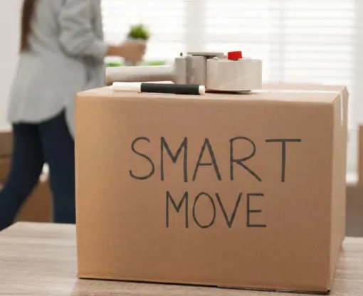 A cardboard moving box has the text "Smart Move" written on it in felt tip. A box sealer is visible on top of the box. A woman moves house plants in the background.