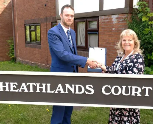 A man and a woman standing in front of a sign that says Heathlands Court, holdng between them a white document