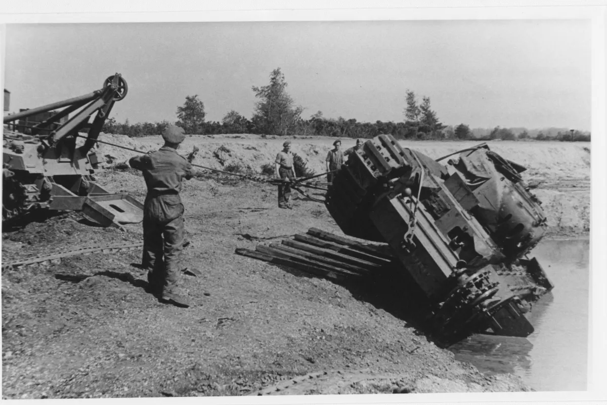 An overturned Churchill tank is being winched out of a pool of muddy water