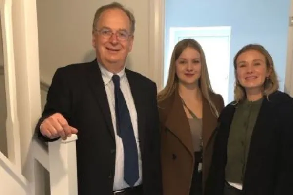 Man standing in the hallway of a house with his right hand on the stairs bannister with two women beside him