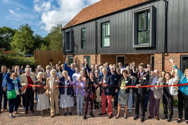 Group of people outside a brick and wood holding a red ribbon which a man is cutting with scissors