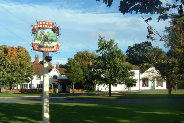 A quaint village scene featuring the "Dog and Partridge" pub sign in the foreground, with a traditional white building surrounded by lush greenery under a clear blue sky.