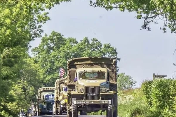 An older military truck leading a procession of classic miliary vehicles along a road with trees and hedges
