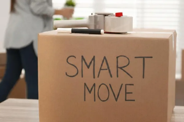 A cardboard moving box has the text "Smart Move" written on it in felt tip. A box sealer is visible on top of the box. A woman moves house plants in the background.