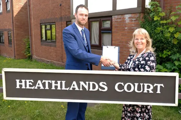 A man and a woman standing in front of a sign that says Heathlands Court, holdng between them a white document