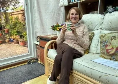 Lady sitting on sofa in her conservatory drinking a cup of tea or coffee smiling